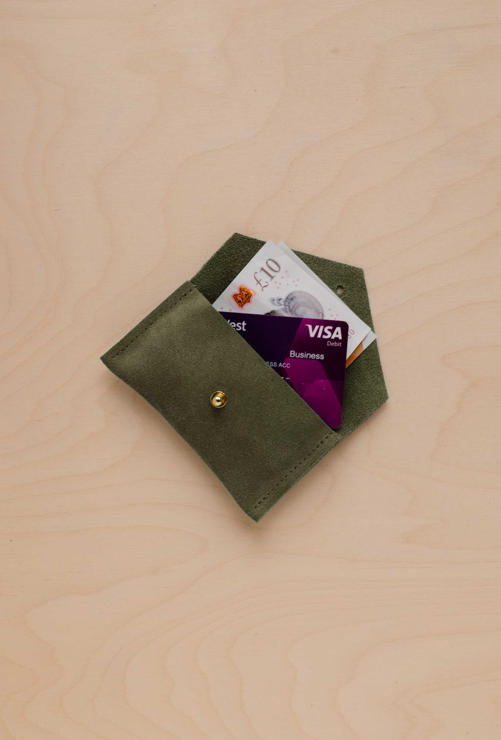 A small soft olive suede pouch with a bank card and bank note nestled inside with a flap and gold button fastening, on a light wood grain surface.