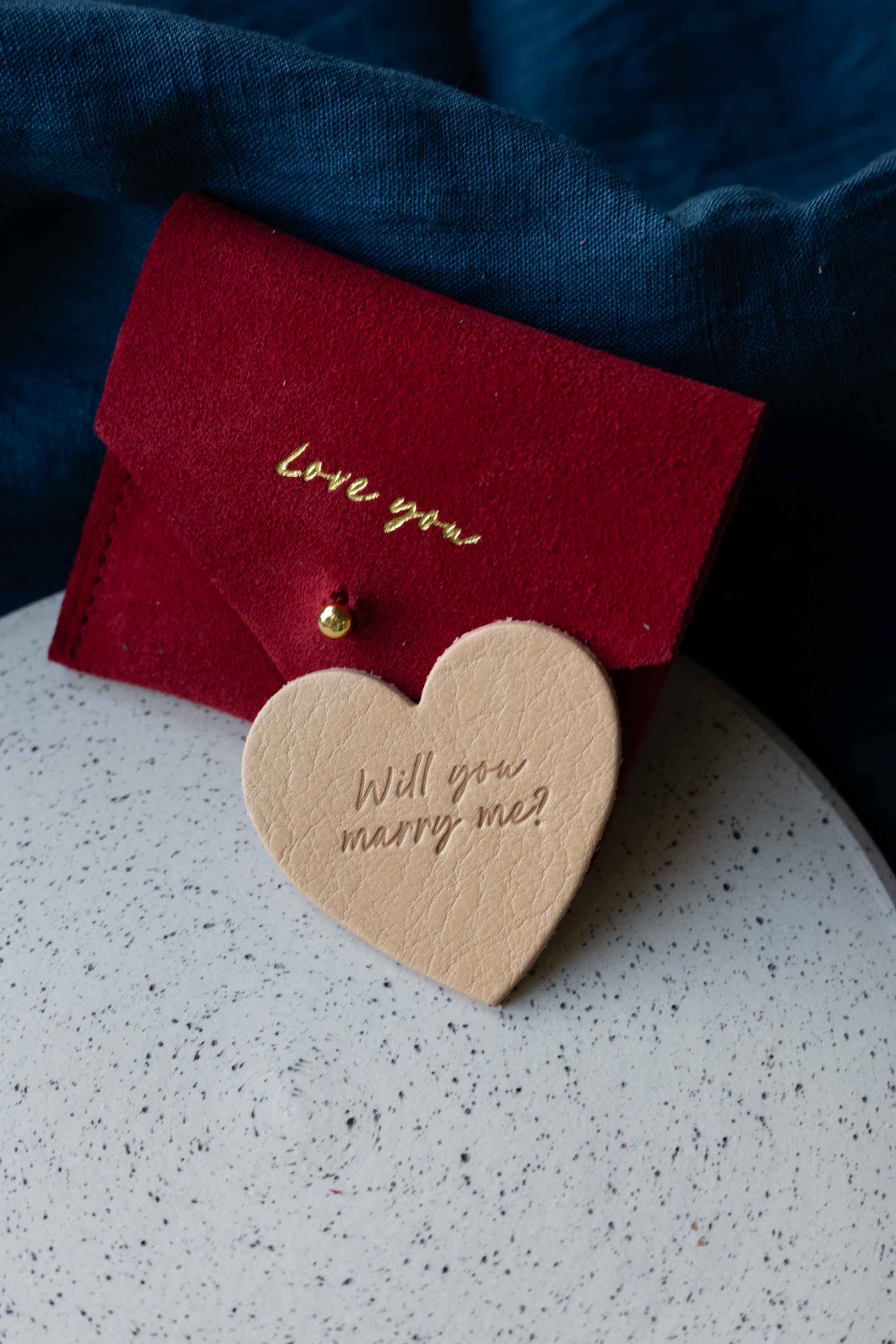 Red card with 'Love you' and heart-shaped wooden tag with 'Will you marry me?' on a speckled surface.