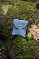 A small dusky blue coloured suede pouch with a gold coloured fastening and flap closure, placed on a natural wooden and moss natural background.