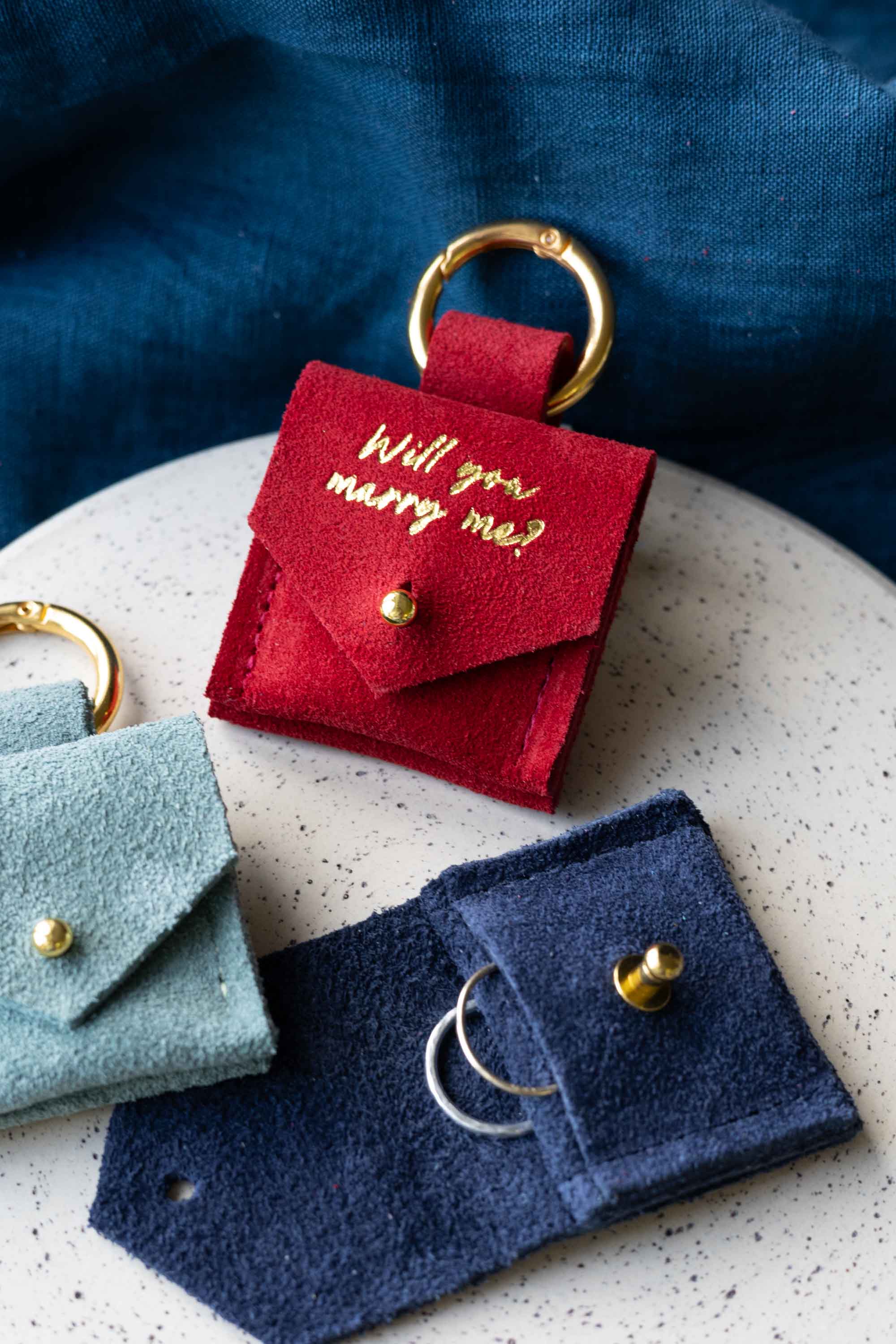 Red, blue, and green suede engagement ring pouches with gold rings on a textured surface. The red pouch features "Will you marry me?".