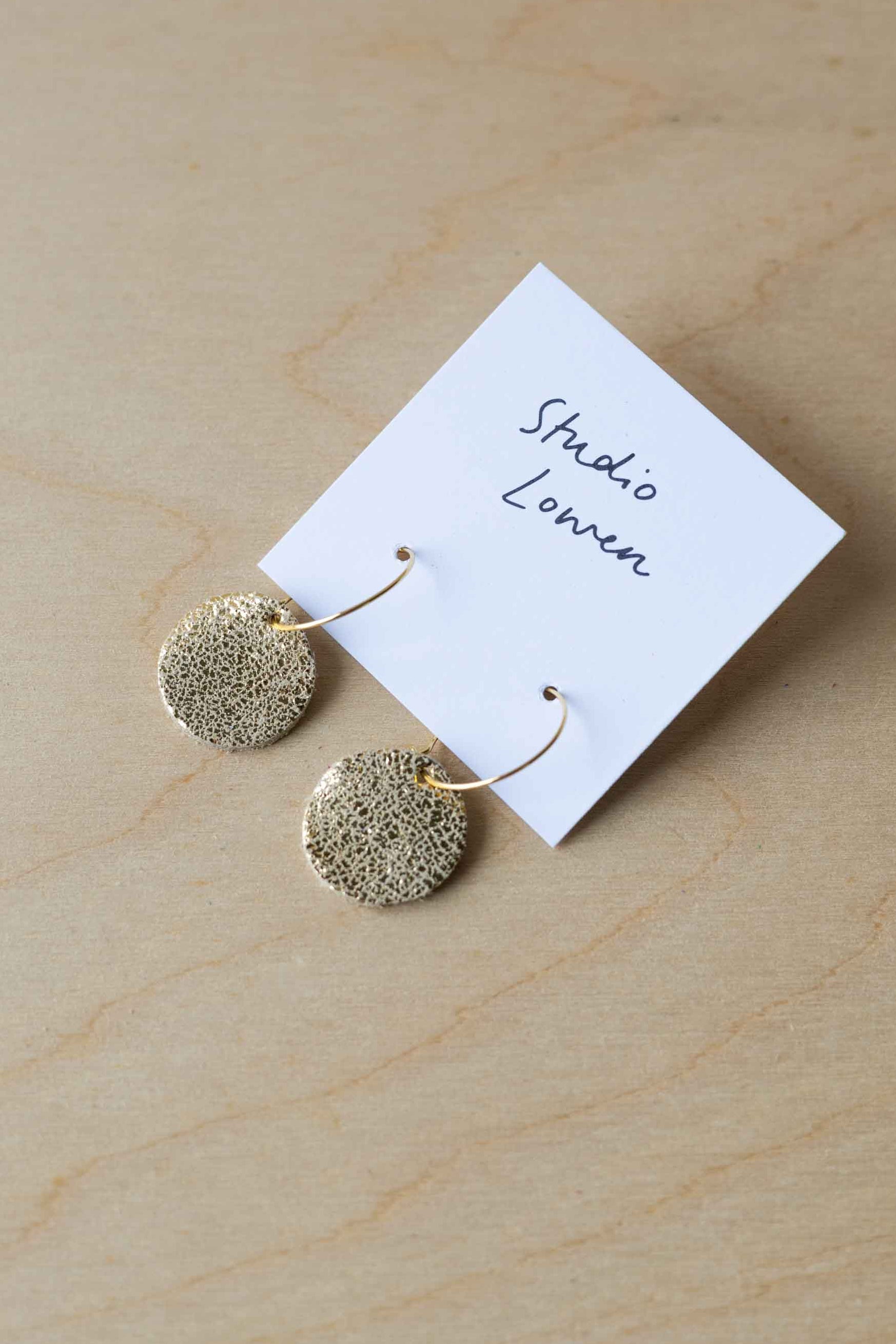 Gold round earrings on a 'Studio Lowen' branded card on a wooden surface