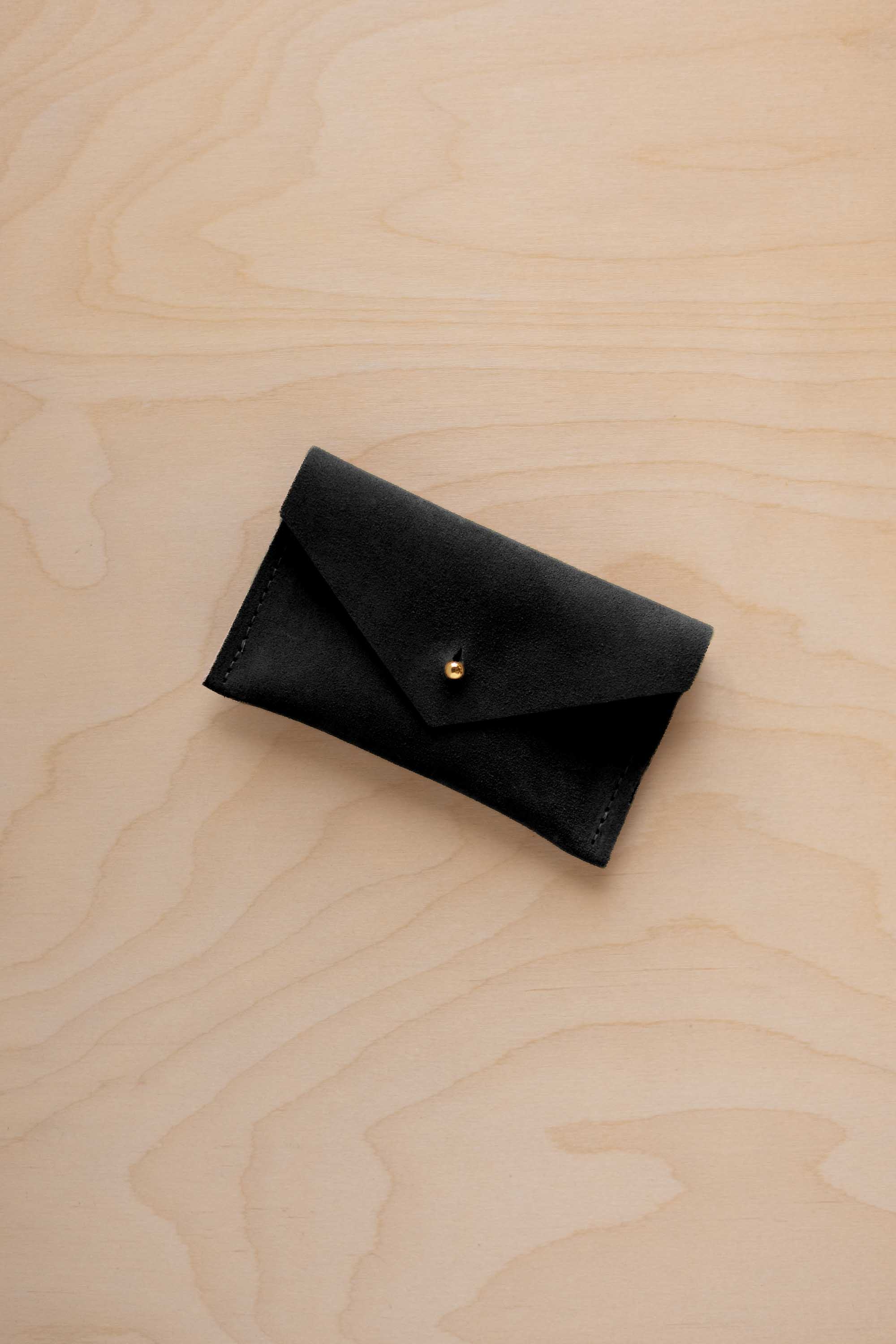 A small black coloured suede pouch with a flap closure and gold coloured fastening, on a light wood grain surface.