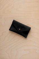 A small black coloured suede pouch with a flap closure and gold coloured fastening, on a light wood grain surface.