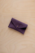 A small blackcurrant coloured suede pouch with a flap closure and gold coloured fastening, on a light wood grain surface.