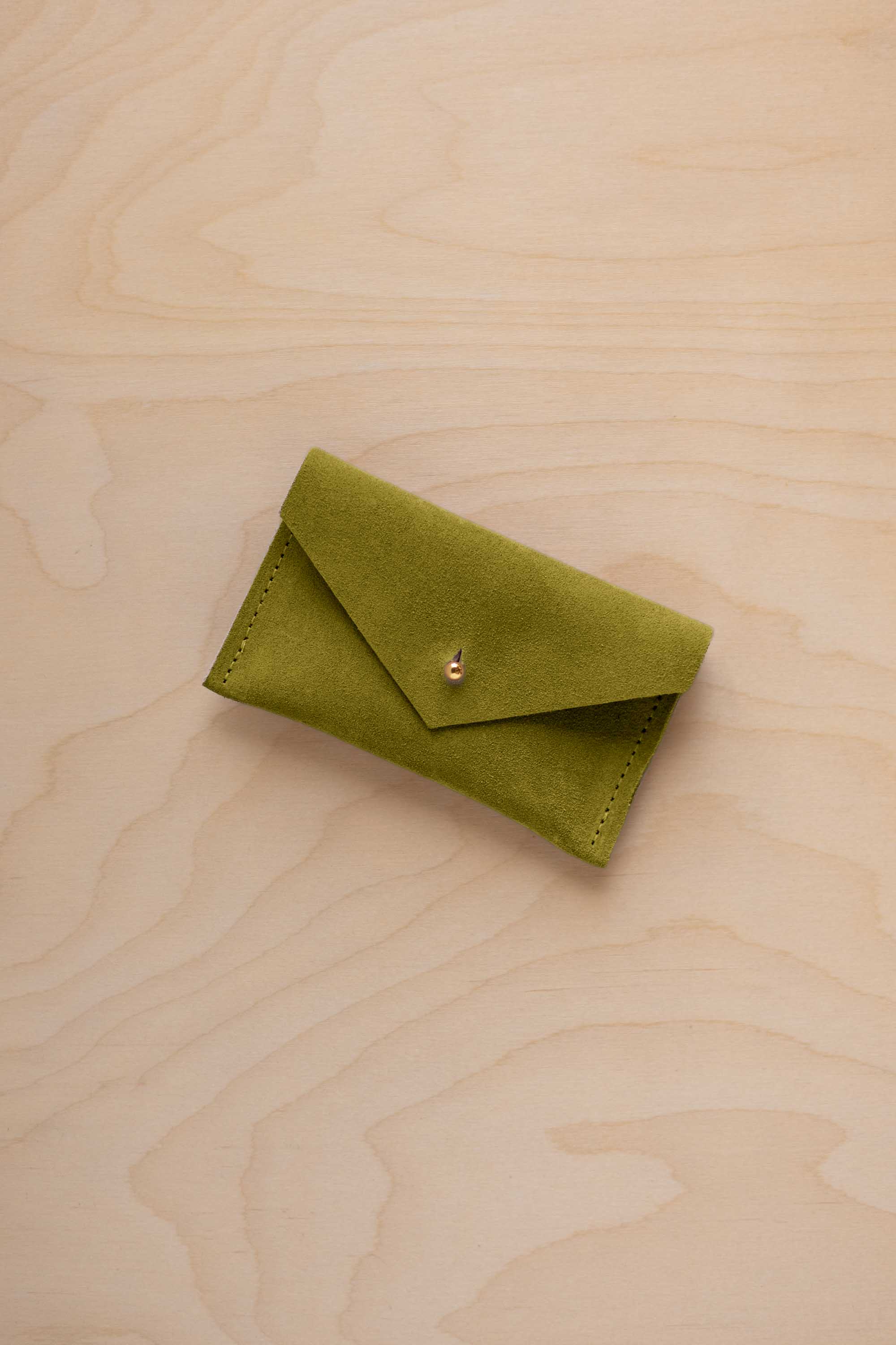 A small chartreuse green coloured suede pouch with a flap closure and gold coloured fastening, on a light wood grain surface.