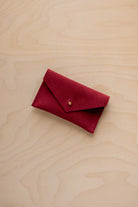 A small crimson red coloured suede pouch with a flap closure and gold coloured fastening, on a light wood grain surface.