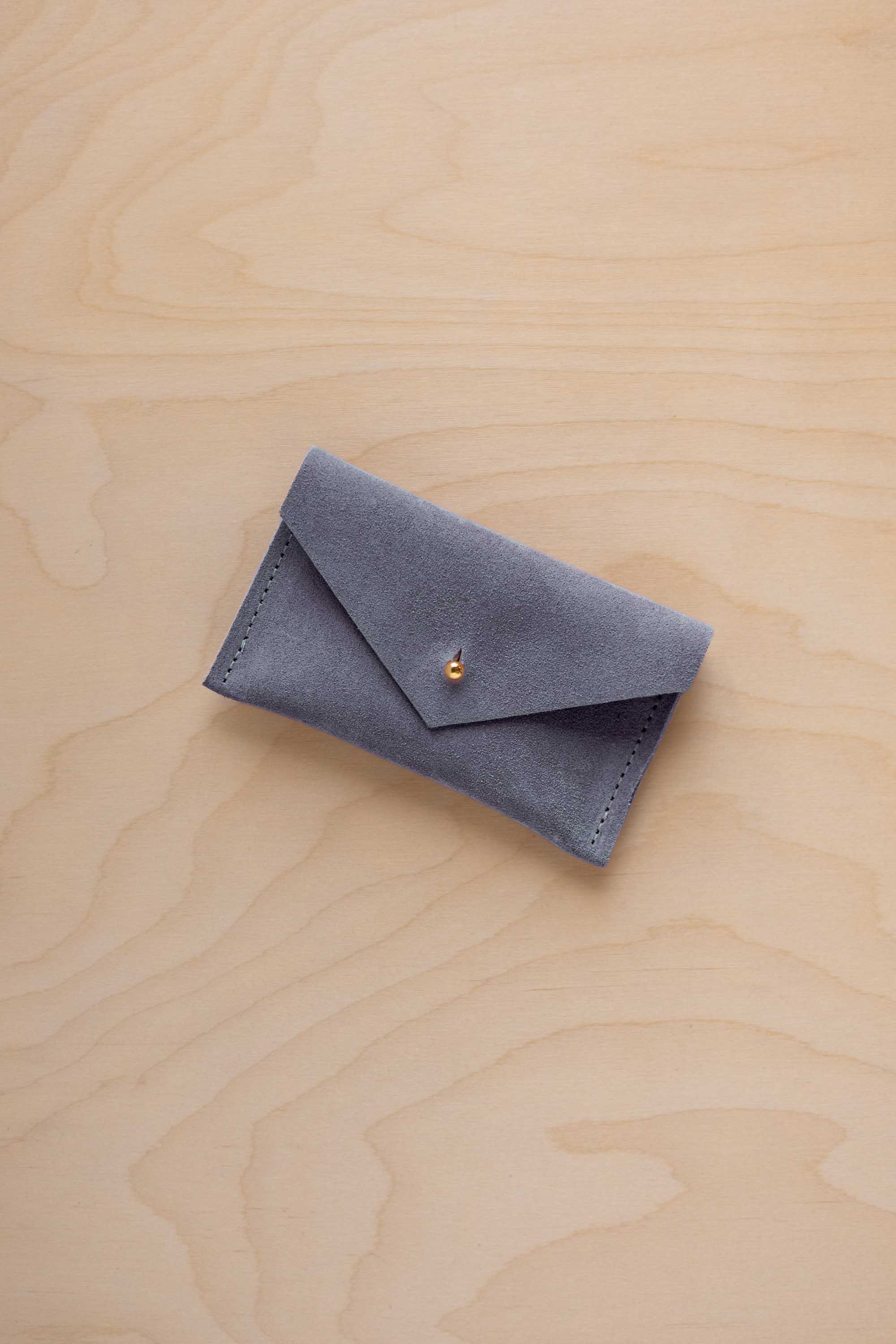 A small dusty blue coloured suede pouch with a flap closure and gold coloured fastening, on a light wood grain surface.