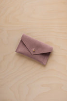 A small dusty mauve coloured suede pouch with a flap closure and gold coloured fastening, on a light wood grain surface.