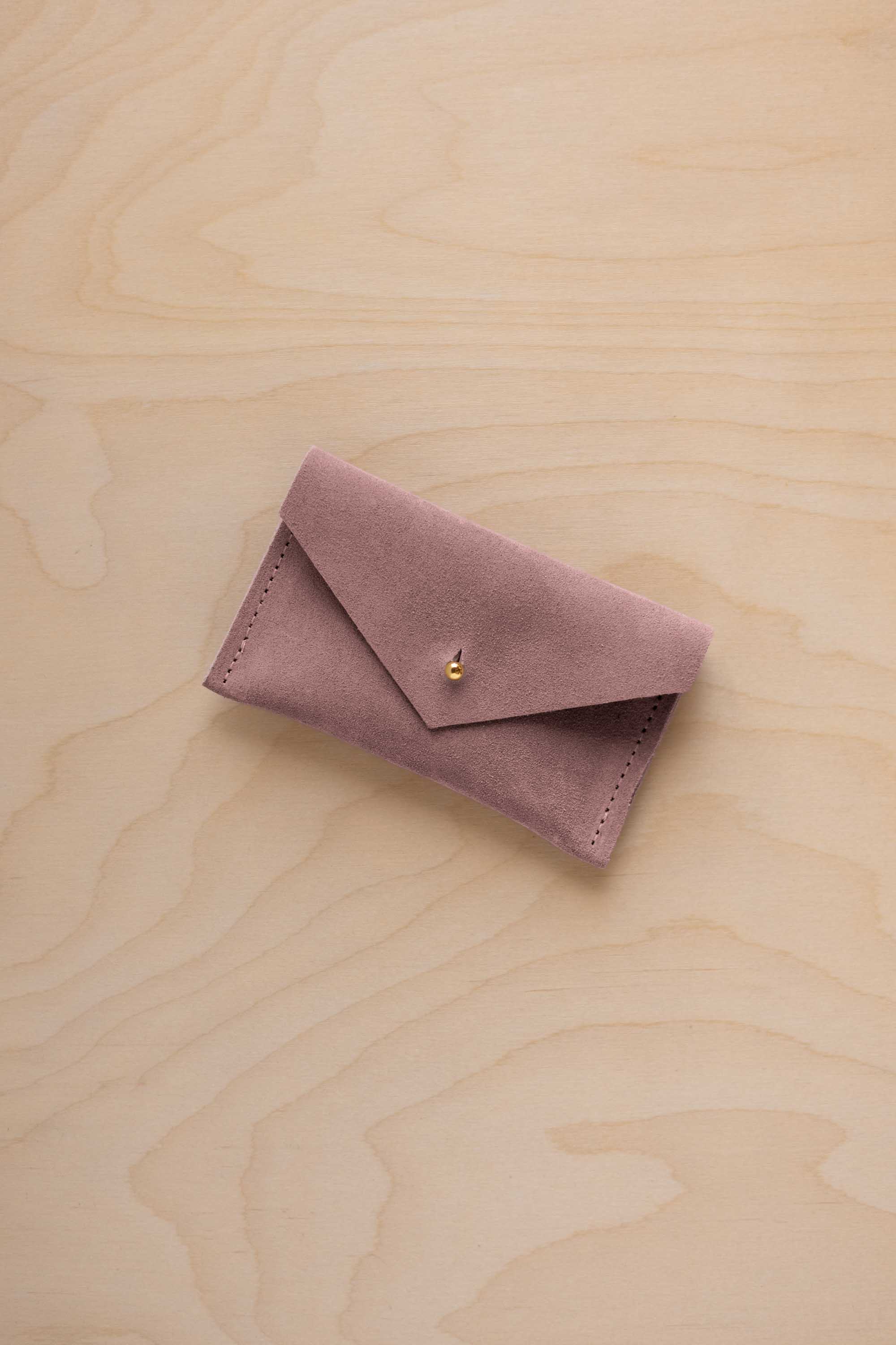 A small dusty mauve coloured suede pouch with a flap closure and gold coloured fastening, on a light wood grain surface.