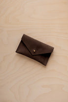 A small espresso brown coloured suede pouch with a flap closure and gold coloured fastening, on a light wood grain surface.