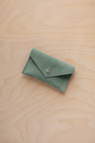 A small eucalyptus green coloured suede pouch with a flap closure and gold coloured fastening, on a light wood grain surface.