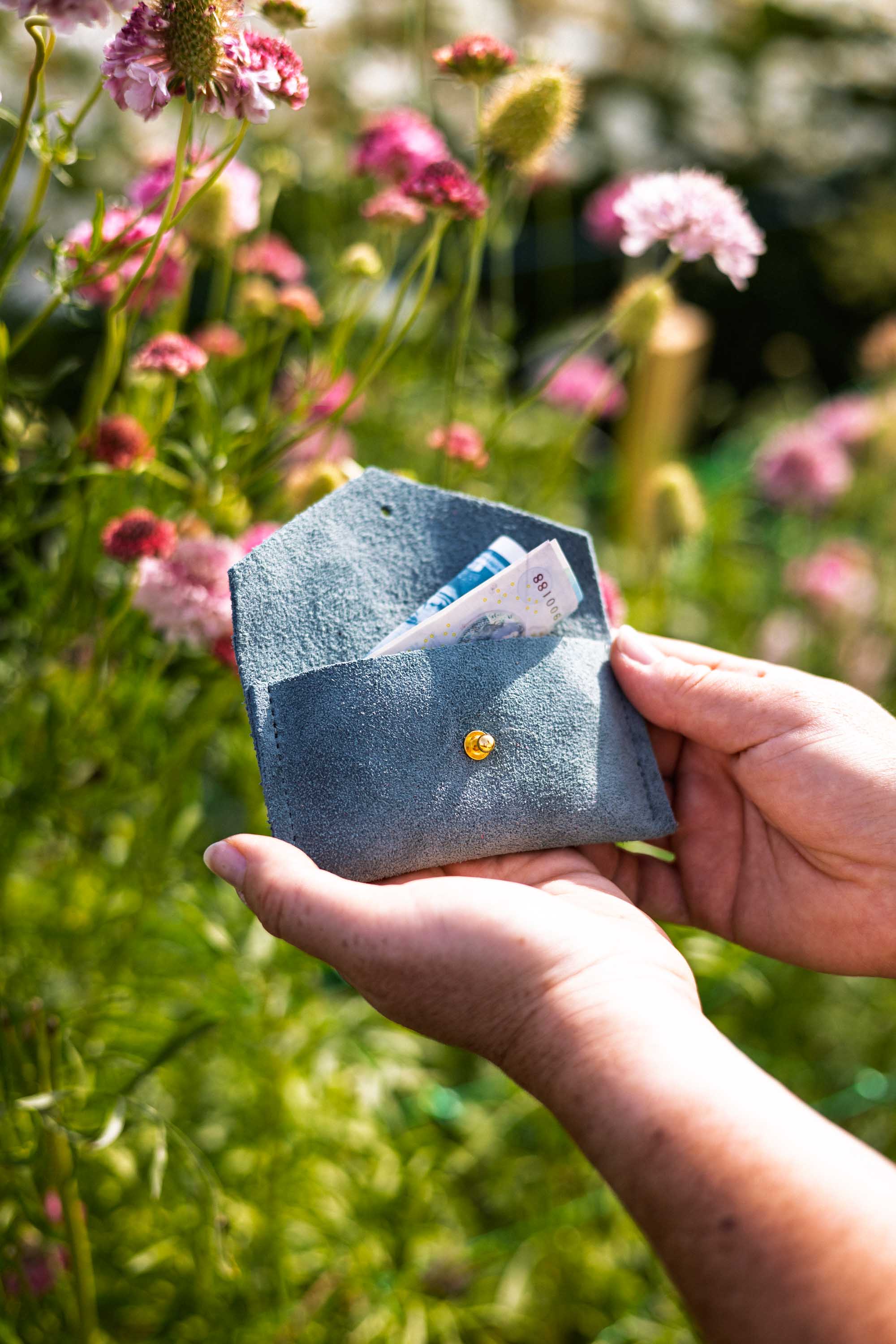 A person's hands holding an open eucalyptus or sage green envelope-style wallet with a gold-tone button, showing a folded money notes inside, set against a blurred background of green foliage and pink wildflowers.