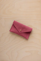 A small faded rose coloured suede pouch with a flap closure and gold coloured fastening, on a light wood grain surface.