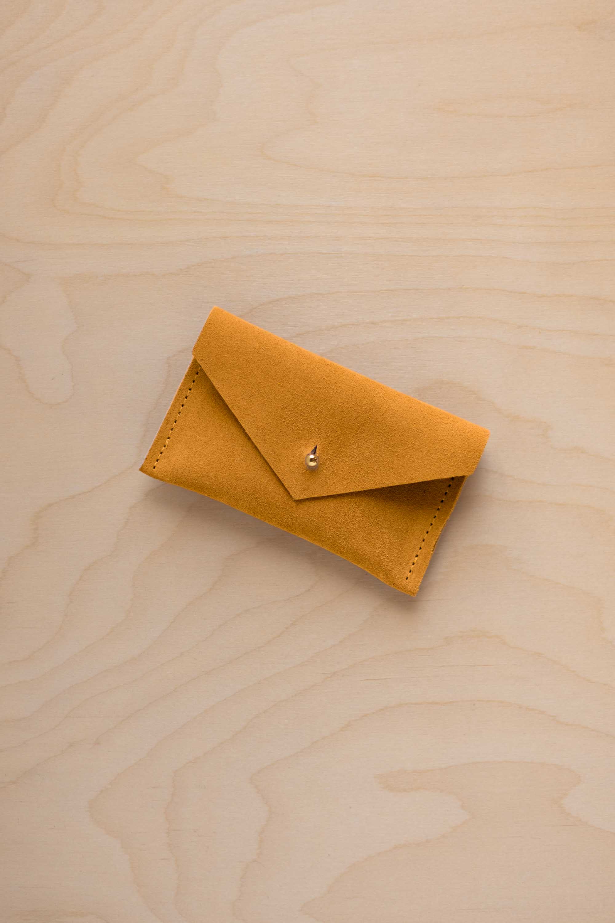 A small golden ochre coloured suede pouch with a flap closure and gold coloured fastening, on a light wood grain surface.