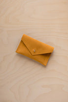 A small golden ochre coloured suede pouch with a flap closure and gold coloured fastening, on a light wood grain surface.