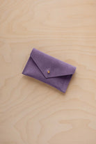 A small lavender coloured suede pouch with a flap closure and gold coloured fastening, on a light wood grain surface.