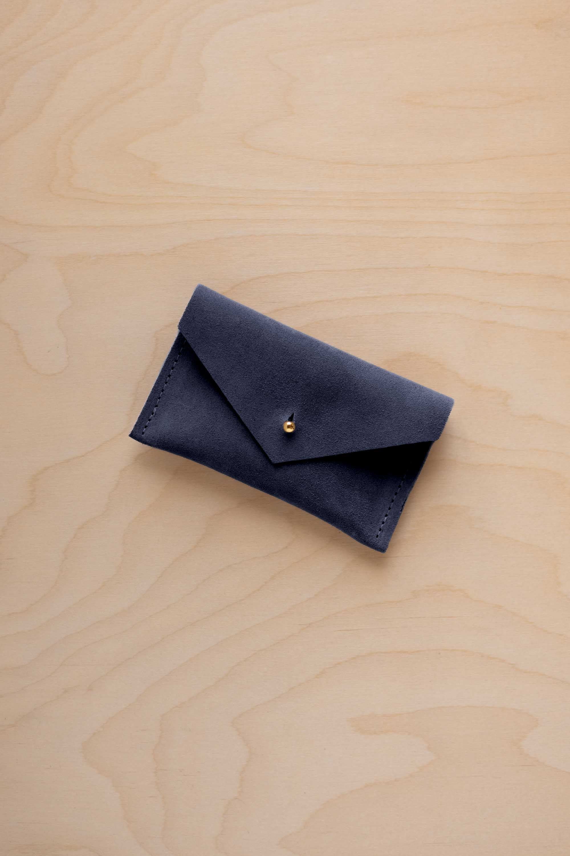 A small midnight navy coloured suede pouch with a flap closure and gold coloured fastening, on a light wood grain surface.