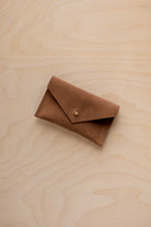 A small mocha brown coloured suede pouch with a flap closure and gold coloured fastening, on a light wood grain surface.