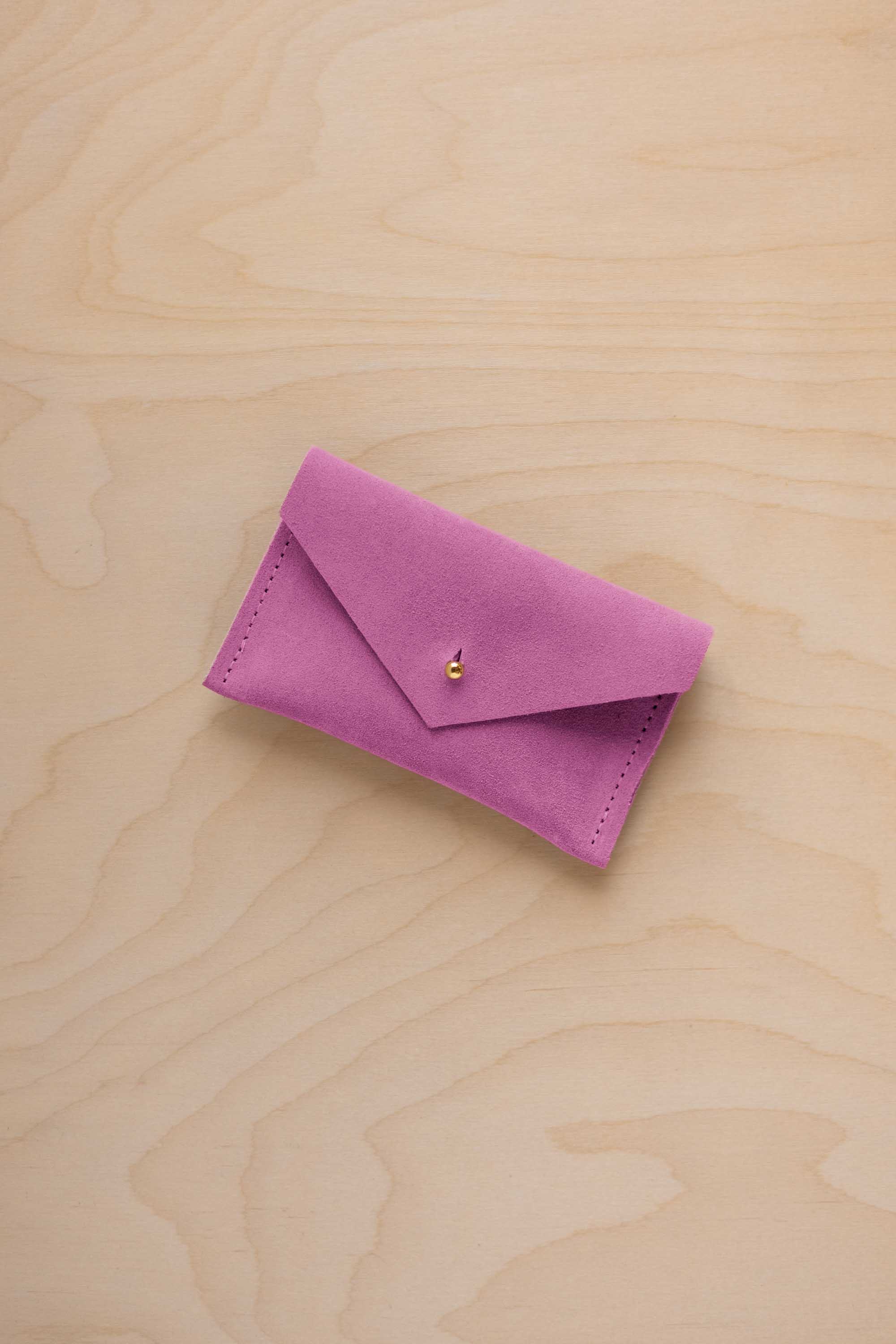 A small peony pink coloured suede pouch with a flap closure and gold coloured fastening, on a light wood grain surface.