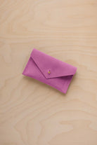 A small peony pink coloured suede pouch with a flap closure and gold coloured fastening, on a light wood grain surface.