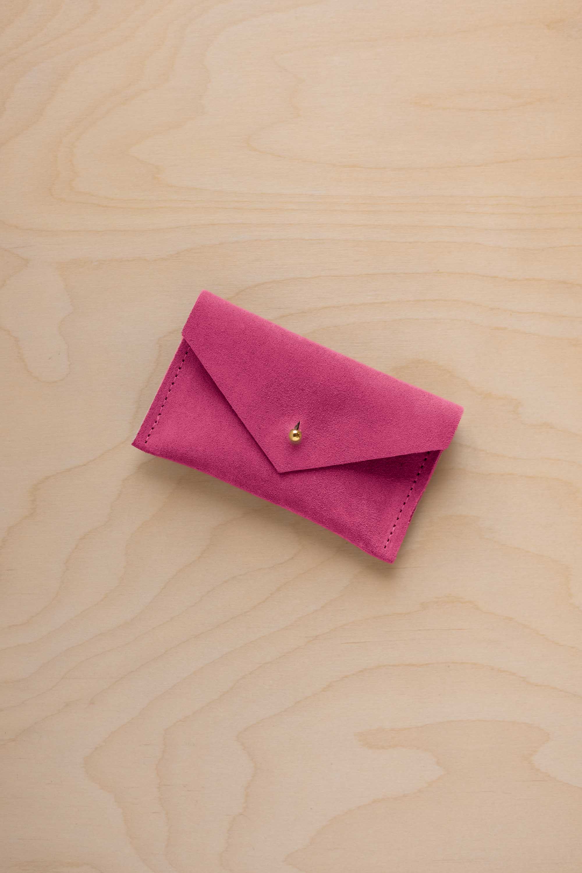 A small raspberry coloured suede pouch with a flap closure and gold coloured fastening, on a light wood grain surface.