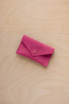 A small raspberry coloured suede pouch with a flap closure and gold coloured fastening, on a light wood grain surface.