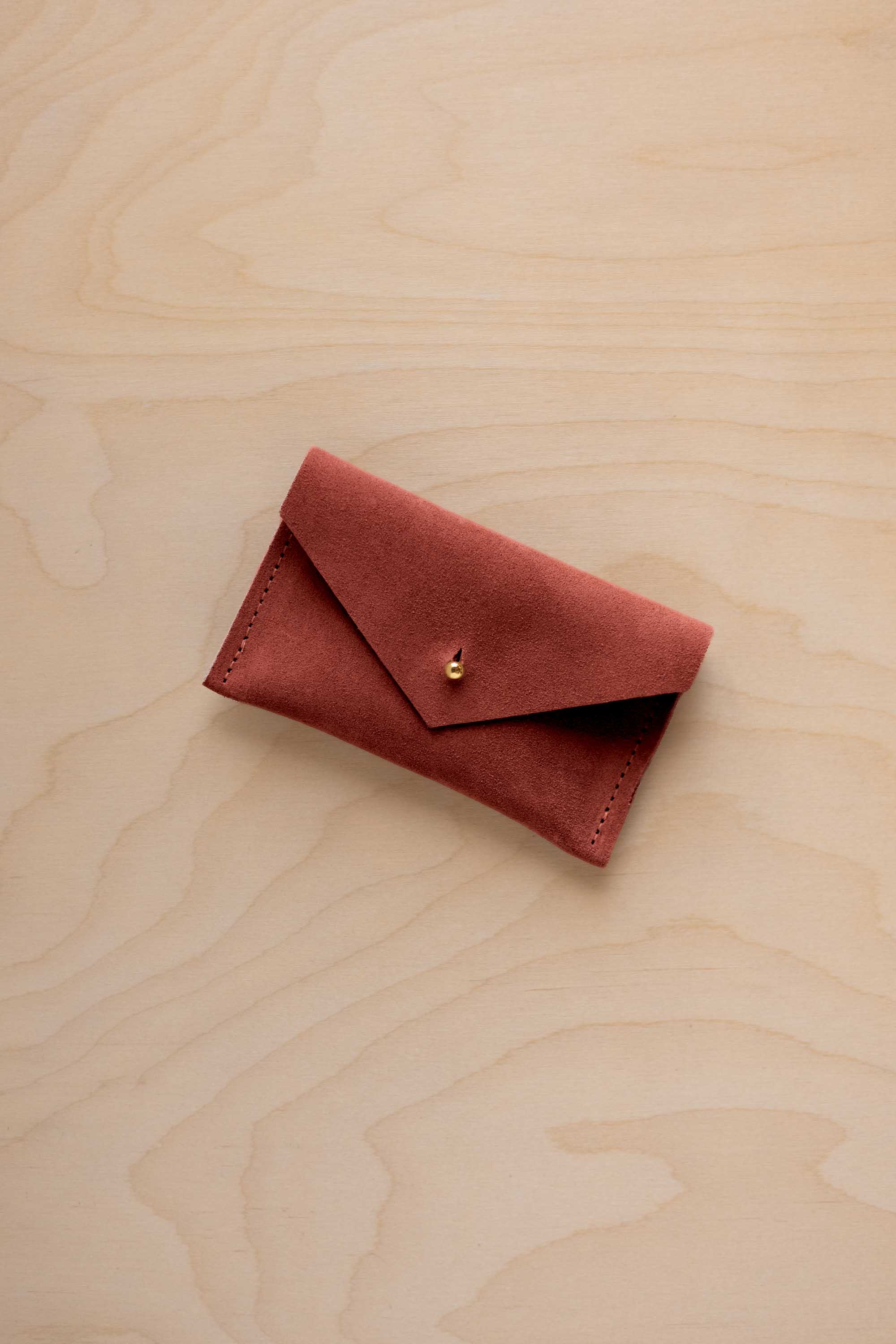 A small rust red coloured suede pouch with a flap closure and gold coloured fastening, on a light wood grain surface.