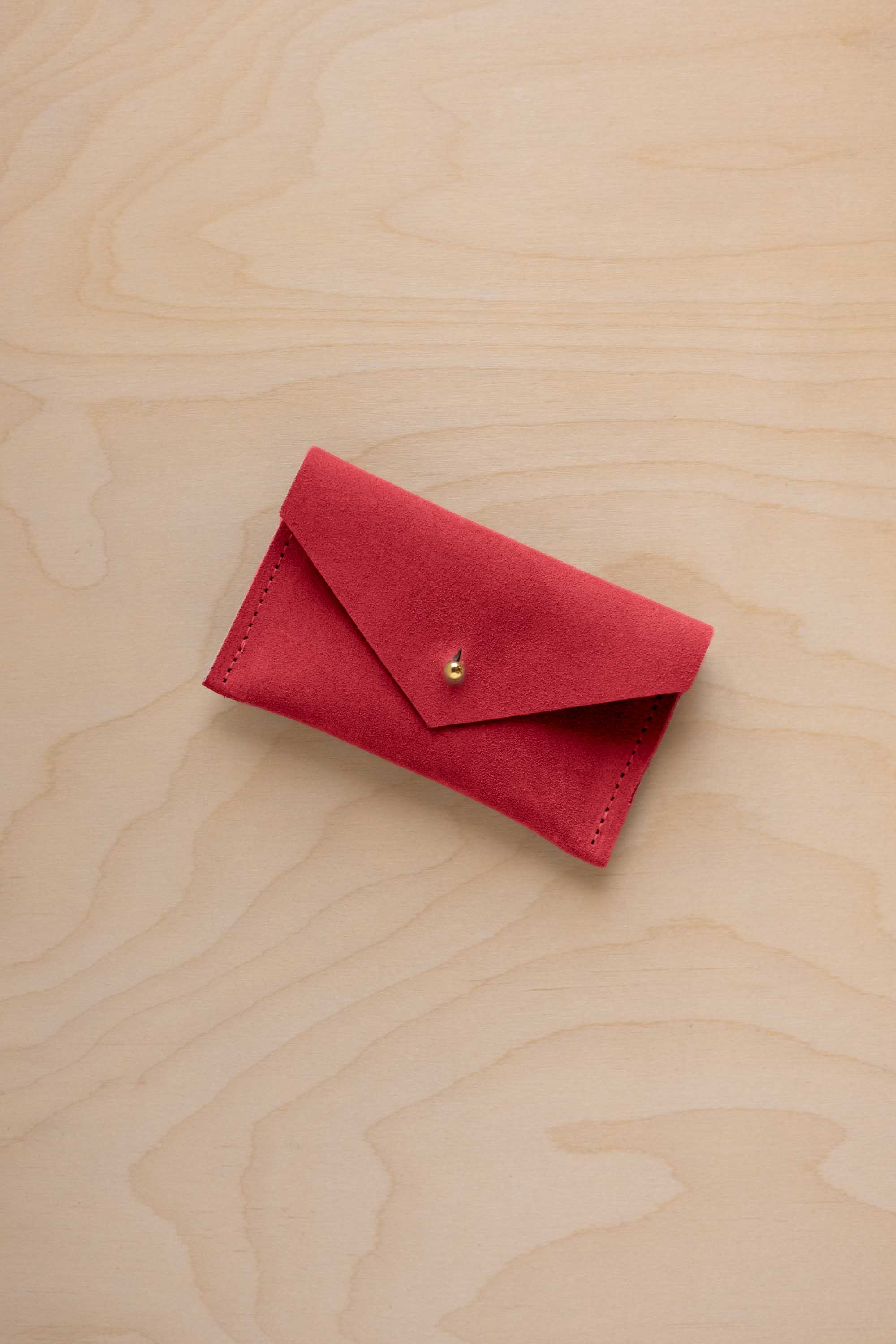 A small scarlet red coloured suede pouch with a flap closure and gold coloured fastening, on a light wood grain surface.