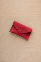 A small scarlet red coloured suede pouch with a flap closure and gold coloured fastening, on a light wood grain surface.