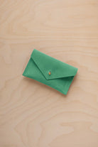A small seafoam green coloured suede pouch with a flap closure and gold coloured fastening, on a light wood grain surface.