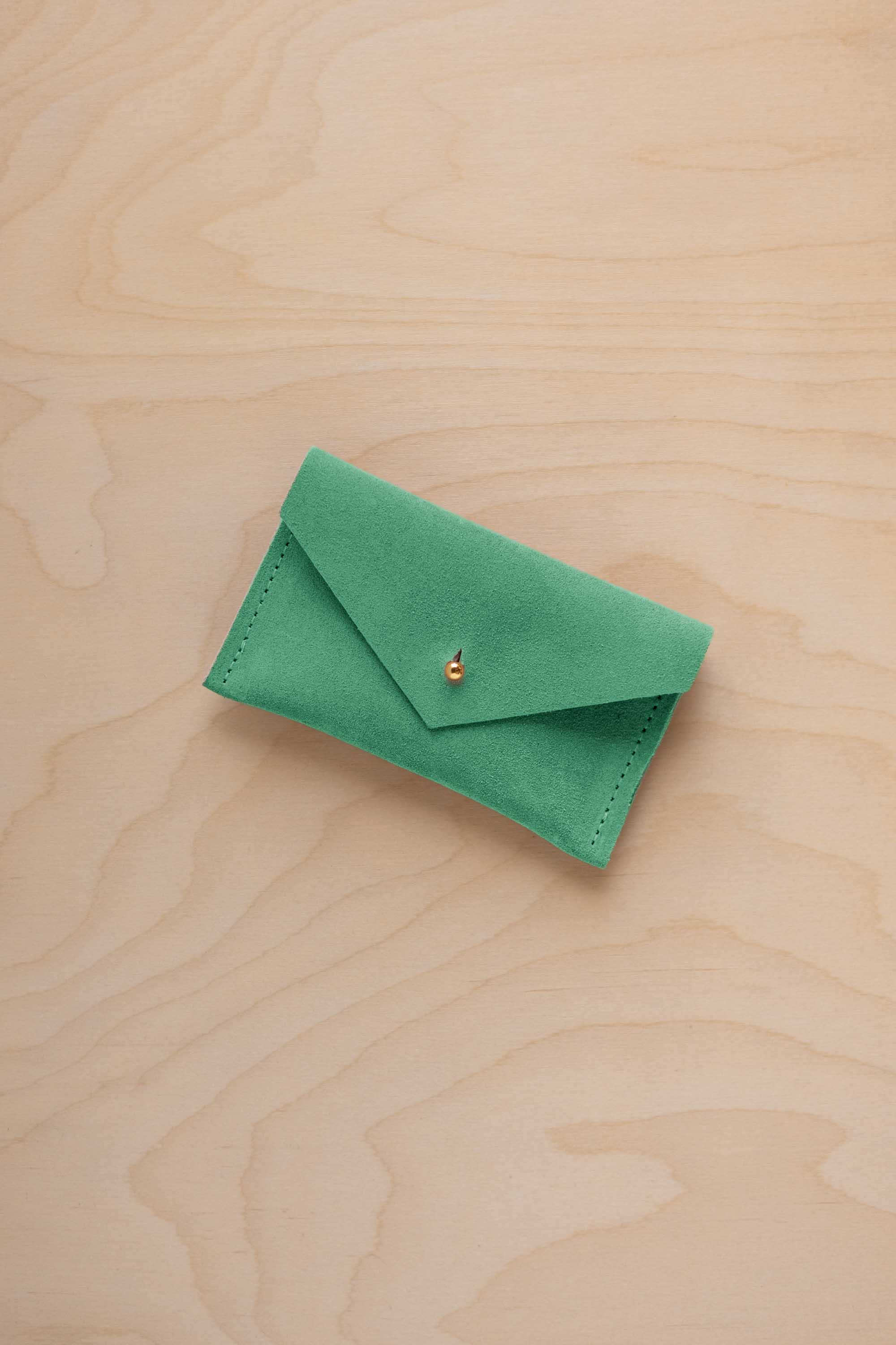 A small seafoam green coloured suede pouch with a flap closure and gold coloured fastening, on a light wood grain surface.
