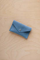 A small sky blue coloured suede pouch with a flap closure and gold coloured fastening, on a light wood grain surface.