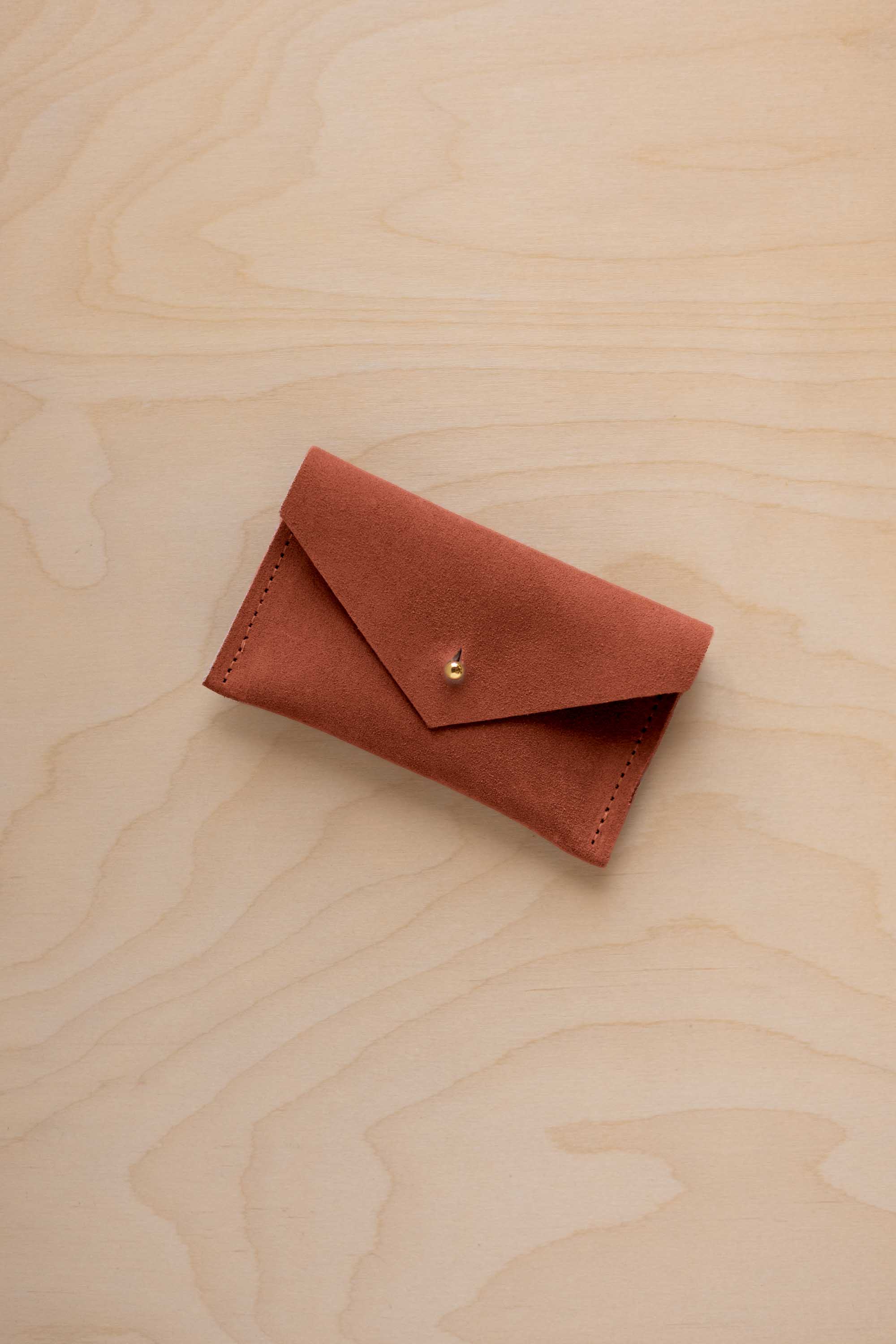 A small terracotta coloured suede pouch with a flap closure and gold coloured fastening, on a light wood grain surface.