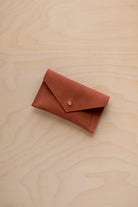A small terracotta coloured suede pouch with a flap closure and gold coloured fastening, on a light wood grain surface.