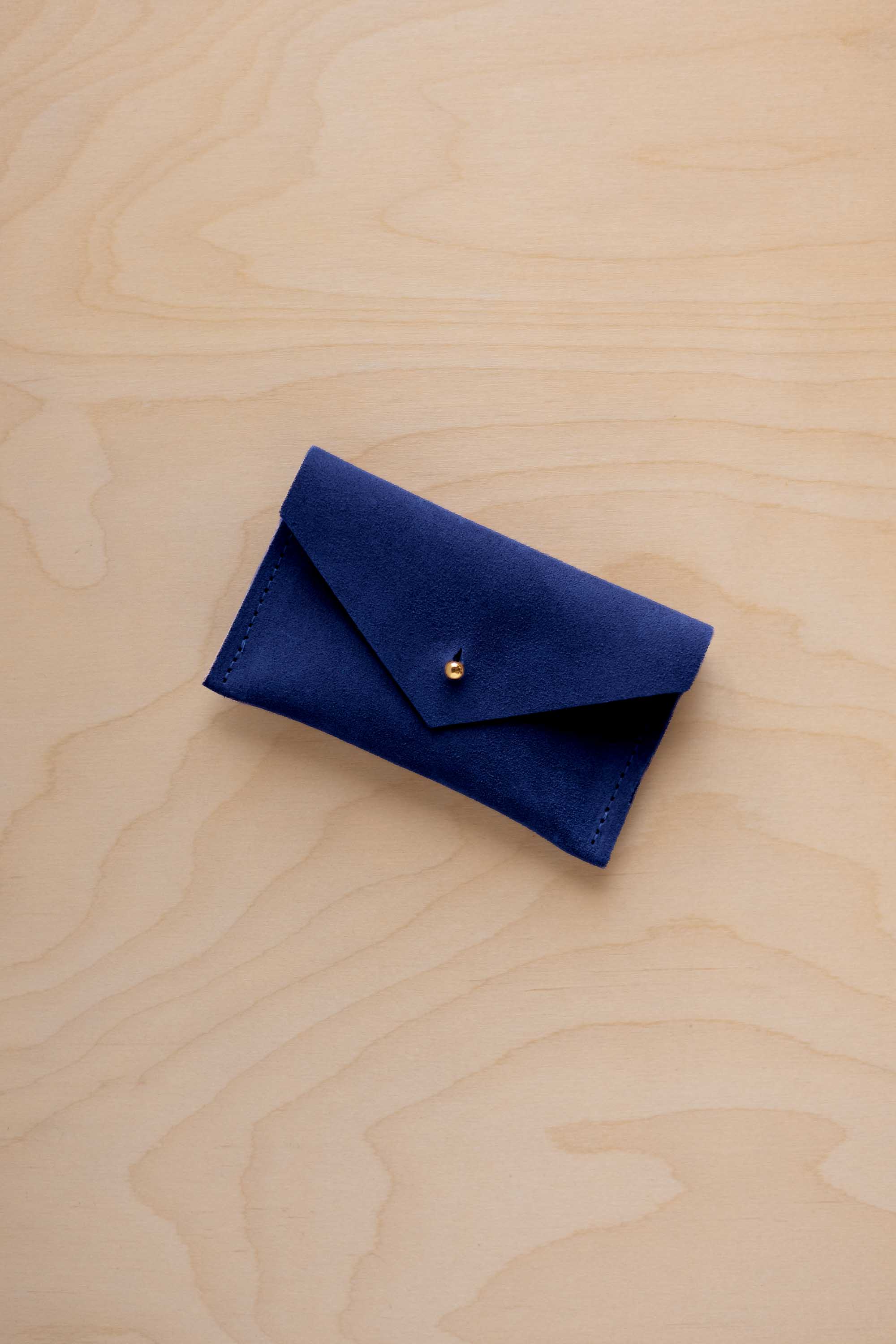 A small ultramarine blue coloured suede pouch with a flap closure and gold coloured fastening, on a light wood grain surface.