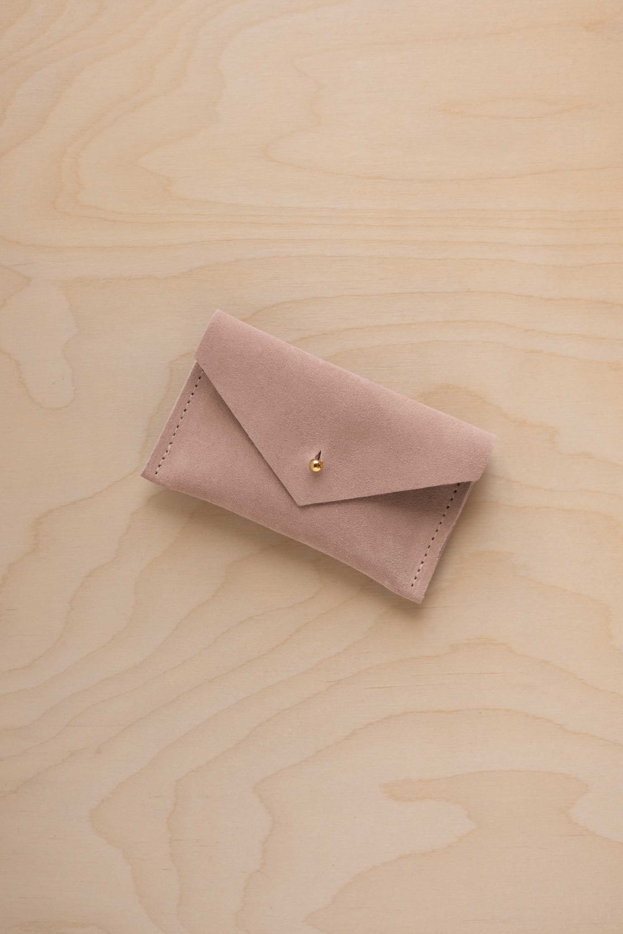 A small warm sand coloured suede pouch with a flap closure and gold button, on a light wood grain surface.
