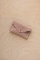 A small warm sand coloured suede pouch with a flap closure and gold button, on a light wood grain surface.