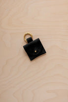 A small black suede pouch bag charm with a gold coloured clip ring fastening, displayed on a light wood grain background.