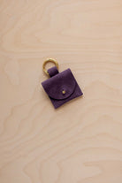 A small blackcurrant dark purple suede pouch bag charm with a gold coloured clip ring fastening, displayed on a light wood grain background.