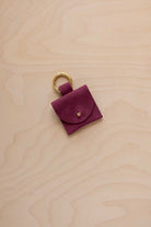 A small burgundy coloured suede pouch bag charm with a gold coloured clip ring fastening, displayed on a light wood grain background.