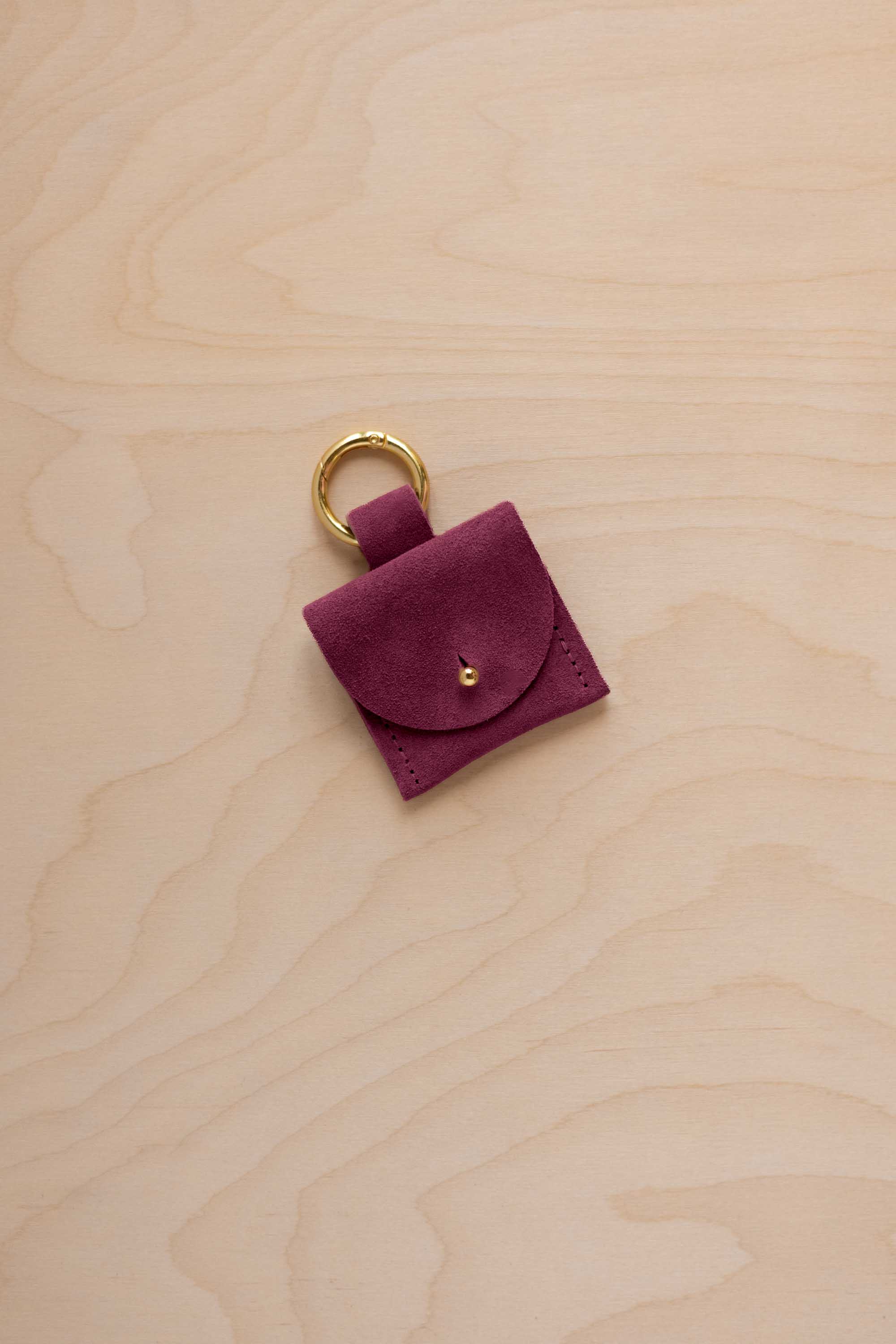 A small burgundy coloured suede pouch bag charm with a gold coloured clip ring fastening, displayed on a light wood grain background.