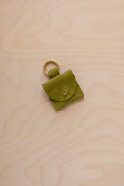 A small chartreuse lime green coloured suede pouch bag charm with a gold coloured clip ring fastening, displayed on a light wood grain background.