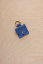 A small cobalt blue coloured suede pouch bag charm with a gold coloured clip ring fastening, displayed on a light wood grain background.