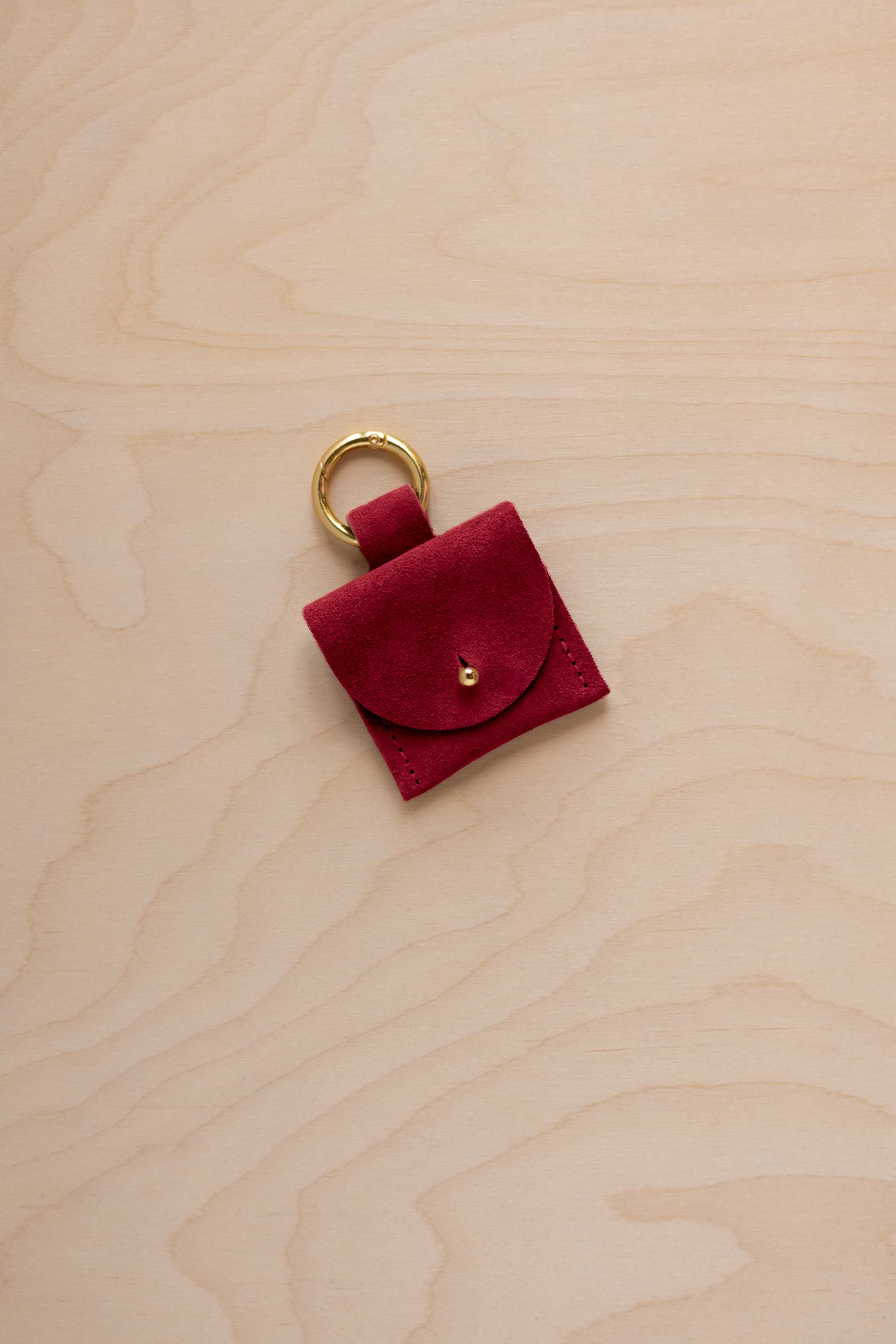 A small crimson red coloured suede pouch bag charm with a gold coloured clip ring fastening, displayed on a light wood grain background.