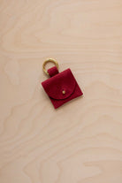 A small crimson red coloured suede pouch bag charm with a gold coloured clip ring fastening, displayed on a light wood grain background.