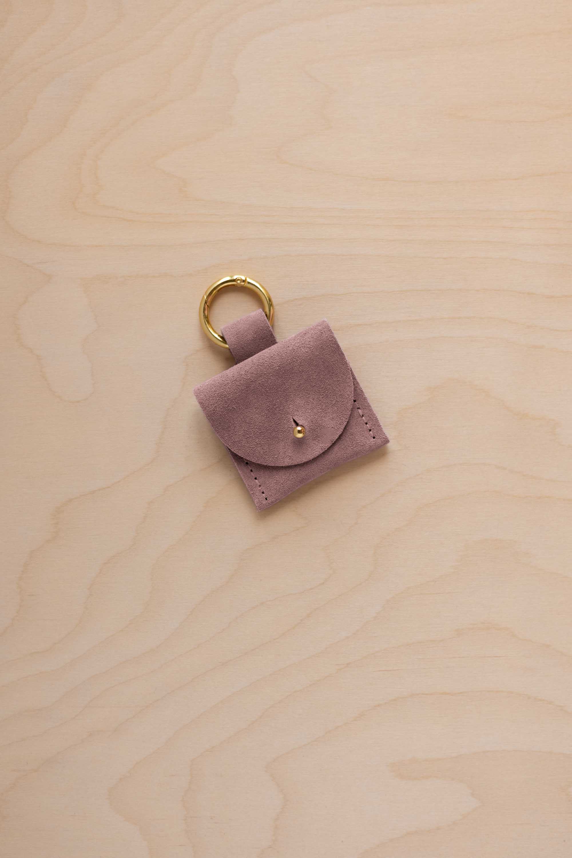 A small dusty mauve suede pouch bag charm with a gold coloured clip ring fastening, displayed on a light wood grain background.