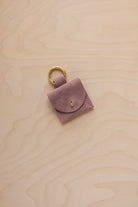 A small dusty mauve suede pouch bag charm with a gold coloured clip ring fastening, displayed on a light wood grain background.