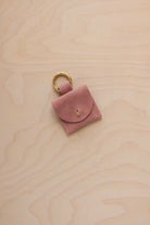 A small dusty rose coloured pale pink suede pouch bag charm with a gold coloured clip ring fastening, displayed on a light wood grain background.