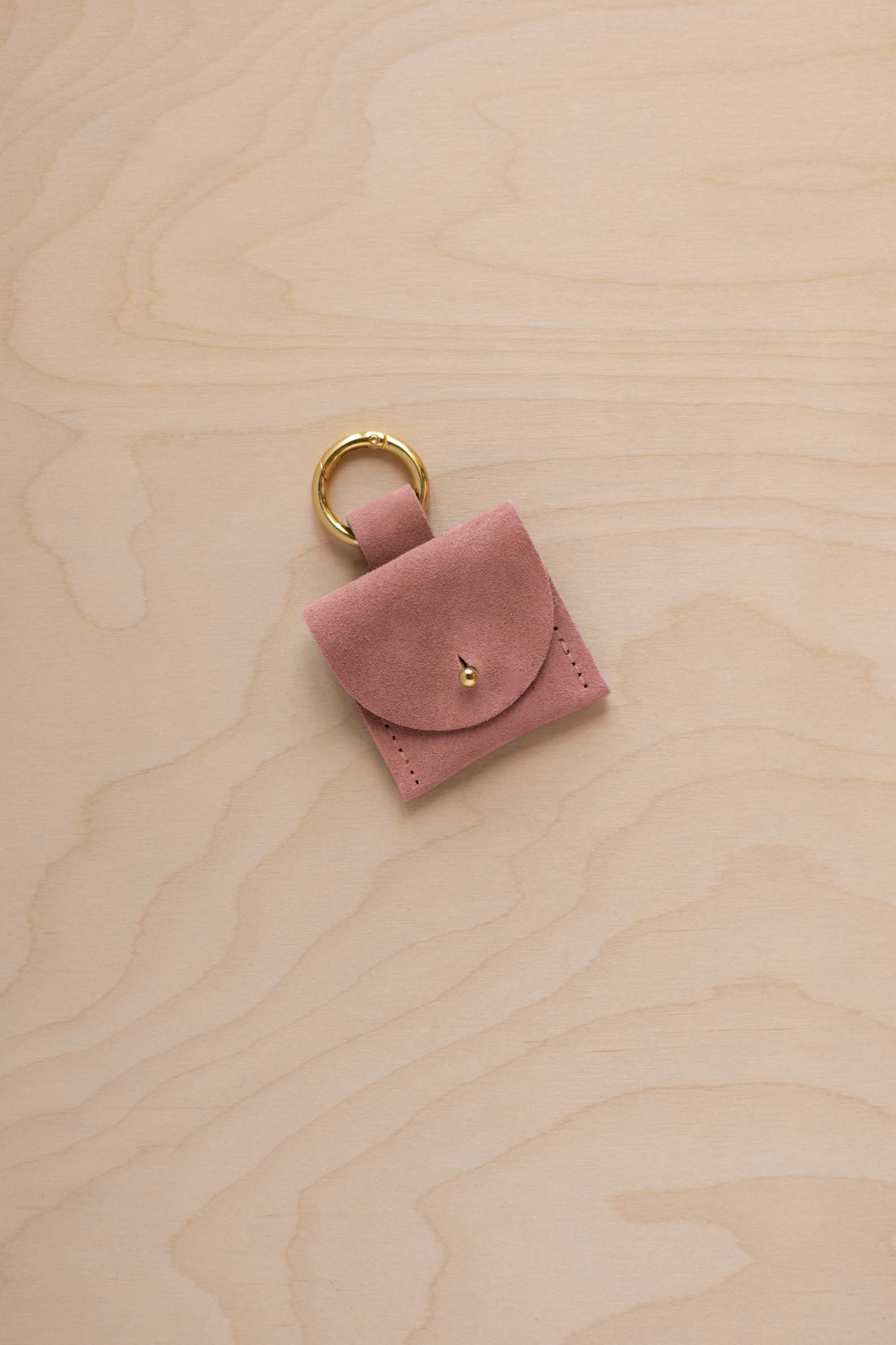 A small dusty rose coloured pale pink suede pouch bag charm with a gold coloured clip ring fastening, displayed on a light wood grain background.