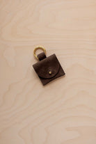 A small espresso brown coloured suede pouch bag charm with a gold coloured clip ring fastening, displayed on a light wood grain background.